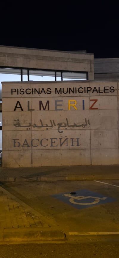 The image shows the exterior of a concrete building at night. The building has large windows on the upper level. The facade of the building features signage in three languages. The top line of text reads "PISCINAS MUNICIPALES" in dark gray letters. Below this, the word "ALMERIZ" is displayed, with each letter a different color: "A" is dark gray, "L" is brown, "M" is dark gray, "E" is yellow, and "RIZ" is red. The third line of text is in Arabic script, "المسابح البلدية" in dark gray. The bottom line of text reads "БАССЕИН" in dark gray Cyrillic letters. In the foreground, there is a parking lot with a paved sidewalk and a curb. A blue square with a white wheelchair symbol is painted on the ground, indicating a disabled parking space. There are no people visible in the image. The scene suggests the entrance to a public swimming pool facility. The city of Huesca, Spain does not have a municipality called Almeriz. Almeriz is not a recognized city or municipality in Spain.