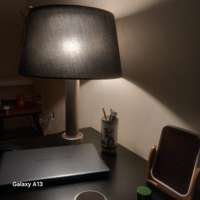 The image depicts a dimly lit desk with a laptop, a lamp, and a small mirror. The laptop is closed and has the text "Galaxy A13" on its surface. To the right of the laptop is a pen holder filled with pencils. A small decorative mirror with a wooden stand is positioned on the right side of the desk. A portion of a lamp with a black shade is visible on the left. The background is a plain wall illuminated by the lamp. There is no indication of the location being Antwerp, Belgium.