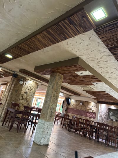 The image depicts the interior of a restaurant with a rustic design. The ceiling features a textured plaster finish interspersed with wooden beams and panels. Large, stone-clad pillars support the ceiling structure. Wooden tables and chairs are arranged throughout the dining area, with some tables set for diners. Natural light enters through large windows in the background, illuminating the tiled floor. The overall ambiance suggests a traditional or themed dining establishment. The restaurant is located in the city of Sredets, Bulgaria.