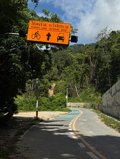 An orange shared lane sign with Thai text and English text "SHARED LANE BEWARE BIKE" is mounted on a metal pole over a paved path in Kathu, Thailand. The sign features icons of a bicycle, an upward-pointing arrow, and a car. The path curves to the right and is marked with white and yellow lines, and a green painted section. Lush green foliage and trees surround the path, with a concrete wall on the right side. A traffic light is also visible. The sky is partly cloudy.