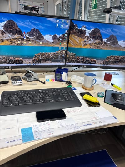 The image shows a cluttered office desk with two large computer monitors displaying a mountainous landscape. In the foreground, a black Logitech keyboard is visible, along with a calculator, a hole punch, and a black smartphone lying on a notepad. There are also various papers, sticky notes, a coffee mug, and office supplies scattered on the desk. A Logitech mouse is positioned on a mousepad to the right. The overall impression is one of a working environment. The specific location in Mannheim, Germany, is not discernible from the visual information provided.