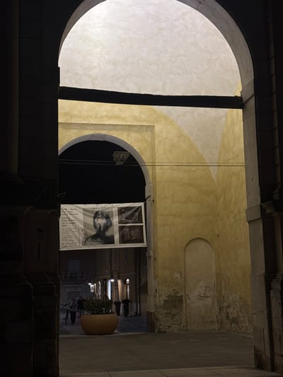 The image depicts an archway leading into a courtyard in Ravenna, Italy. A banner advertising an exhibition at the MAR - Museo d'Arte della città di Ravenna, Galleria Paolo Roversi, is hung under a second arch. The banner features a portrait and other graphic elements. In the courtyard, two individuals are walking, one pushing a bicycle. A large planter with greenery is centrally located. Streetlights illuminate the scene. The architecture suggests a historic urban environment, consistent with Ravenna's character.