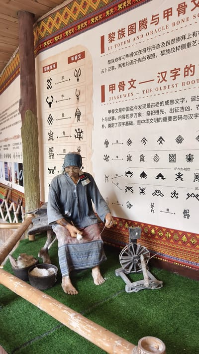 Within an indoor exhibit in Jiyang District, China, an elderly female mannequin is positioned, depicting the act of hand-spinning. The mannequin is barefoot, with dark skin, a blue headscarf, a loose blue top, and a long, patterned skirt featuring red, blue, and white stripes and geometric designs. Traditional tattoo-like markings are visible on her forearms and lower legs. She is seated on a low, rustic wooden bench.
Her hands are posed actively, with the right hand holding a small, spherical white object (likely spun fiber or a drop spindle), and the left hand drawing out unspun material. A thin thread connects her hands to a dark, spoked spinning wheel situated on the artificial green grass to her right. To her left, two dark, woven baskets rest on the artificial grass; one contains fluffy, raw white fiber, and the other holds processed white fiber. A large, light-colored bamboo pole lies horizontally in the foreground, and another leans diagonally behind the baskets.
The background is dominated by a large informational panel with Chinese text and English translations, including "LI TOTEM AND ORACLE BONE SCRIPT" and "JIAGUWEN - THE OLDEST ROOT". The panel displays various symbols and pictograms, connected by arrows, illustrating representations of animals such as sheep, ox, fish, turtle, and deer. The panel's top and bottom edges feature decorative borders with repetitive geometric patterns in red, yellow, and black. A thick wooden support beam is visible on the left, and the ceiling exhibits exposed wooden beams and a patterned border.