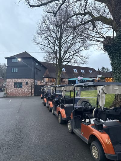 This image depicts an outdoor scene at the Old Thorns Manor Hotel & Golf Course in Griggs Green, Liphook, East Hampshire, United Kingdom, under an overcast sky. In the foreground, a line of at least seven orange and black golf carts with light-colored fabric roofs is parked on an asphalt surface. Visible cart numbers include 18, 22, and 4. To the left, a multi-story building features a red brick lower section and dark grey horizontal siding on its upper floors, with several windows and an illuminated exterior floodlight. A dark gate or door is visible adjacent to this building. In the background, a larger, multi-roofed building displays a prominent light blue sign with "Oties" in white script and features dormer windows on its dark roof. Green grass is visible between the golf carts and this background building. Bare deciduous trees are present, including a large one centrally positioned and another on the far right covered in ivy. No individuals are visible.