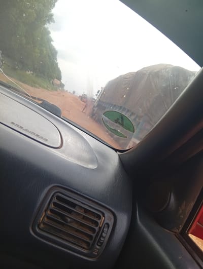The image is taken from inside a vehicle looking forward. The dashboard is visible in the foreground with an air vent and "SRS AIRBAG" text. Through the windshield, a dirt road is visible with trees lining the left side. In the distance, a large truck covered with a tarp is stopped on the road. The sky is overcast. The location is specified as Ndavangva, Nigeria.