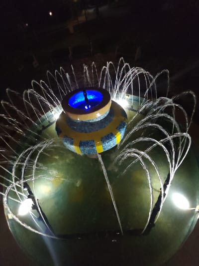 The image depicts an illuminated water fountain at night in Veliki Preslav, Bulgaria. The central feature is a tiered structure, cylindrical in form, adorned with blue and yellow mosaic tiles arranged in a band pattern. The top-most tier contains a concave basin emitting a bright blue light from within, with a small central water feature. Numerous thin water jets emanate in an arc pattern from the perimeter of the central tiled structure and also from the outer basin, creating a circular display. The main fountain basin is filled with dark green water, illuminated by several distinct white lights positioned around its outer edge, which highlight the ascending water streams. The surrounding environment is dark, with faint outlines of indistinct structures or benches visible in the distant background, suggesting an outdoor public space. No individuals or specific identifying landmarks beyond the fountain are discernable.