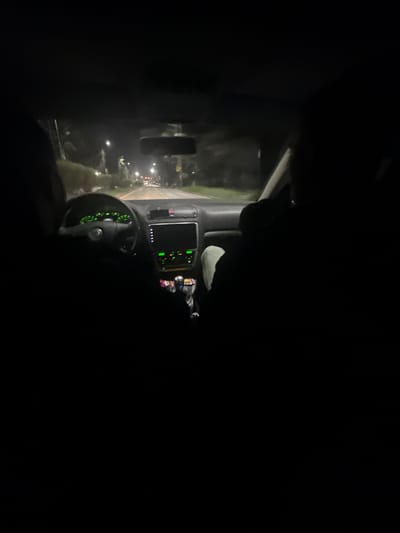 The image shows the interior of a car at night. The dashboard and steering wheel are visible, illuminated by green lights. Through the windshield, there is a blurred view of a road with streetlights. The specific location within Pernik, Bulgaria, cannot be determined from the image.
