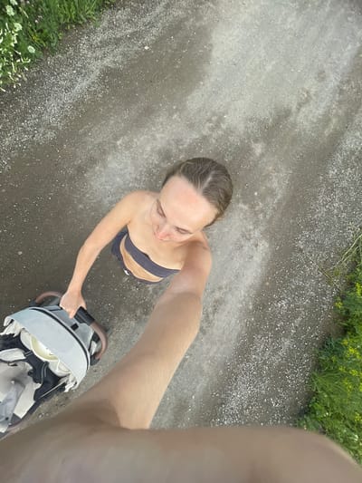 An overhead shot captures a woman from the shoulders up, pushing a baby stroller along a gravel path. She is looking down and to her right, with her hair pulled back. She wears a dark-colored bandeau top and dark shorts. The stroller is light gray and white. To the left of the path, there is green grass with some small pink and white flowers. To the right of the path, there is more green vegetation with yellow flowers. The ground is predominantly composed of gray gravel with some larger stones. The scene appears to be outdoors, possibly in a park or a less-developed area, given the natural vegetation and gravel path. The photograph is taken from a high angle, looking down at the woman and the stroller. The location is identified as Podshivalovo, Russia.