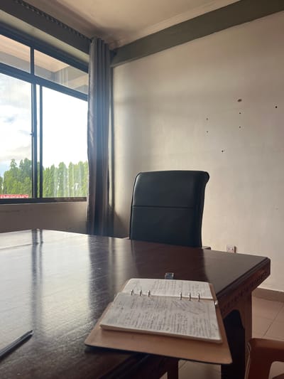 The image shows an interior scene in Picha Ya Ndege, Tanzania. A black leather office chair is positioned at a dark wooden desk. On the desk, there's an open notebook with handwritten notes. A pen is lying on the desk. A window, partially obscured by grey curtains, offers a view of trees outside. The ceiling is white, and there are some visible holes or imperfections. An electrical outlet is visible on the wall.