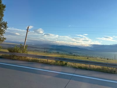 A wide-angle shot captures a rural landscape with rolling hills under a bright blue sky dotted with scattered clouds. In the foreground, a grey asphalt road with a white dividing line runs diagonally across the frame, partially obscured by the shadow of an unseen object. A metal guardrail runs parallel to the road, separating it from a grassy embankment. Beyond the embankment, a lush green field stretches out, patterned with darker green patches that could be crops or trees. A lone wooden utility pole stands tall in the middle ground, its wires extending across the frame. In the far distance, a range of hazy, blue-toned mountains forms the horizon. The overall scene depicts a serene, natural environment bathed in natural light, suggesting an outdoor setting in a rural area. The image does not contain any people or animals.
