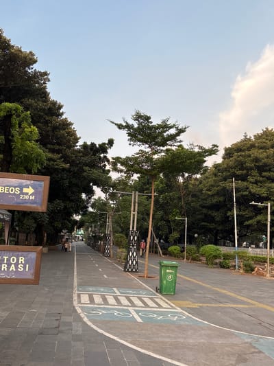 The image depicts a paved pathway lined with trees, likely in a park or urban green space. On the left, a sign with blue lettering and an arrow pointing right reads "BEOS 230 M." Below it, another sign partially visible says "TOR ERASI." A green trash bin is situated on the right side of the pathway. The pathway is marked with white lines and symbols indicating a bike lane. Several trees, some tall and slender and others with dense foliage, create a canopy overhead. In the background, people can be seen walking along the path. The sky above is a pale blue with a few wispy clouds. The overall scene suggests a well-maintained outdoor area in a city. This location is in the city of West Jakarta, Indonesia.