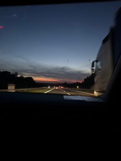 The image depicts a view from inside a vehicle traveling on a highway at dusk. The sky is a deep blue transitioning to orange and pink hues near the horizon, indicating sunset. Silhouetted trees line the sides of the highway, with some distant lights visible. Several vehicles, including cars and a large truck, are on the road, with their headlights on. The road itself is clearly marked with white lines. The foreground is dark, suggesting the interior of the car, with part of the windshield and dashboard visible. The image does not contain information that would allow for identification of the specific location within Mysłowice, Poland.