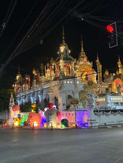 The image captures Wat Ming Muang, a Thai Buddhist temple, at night in Chiang Rai City Municipality, Thailand. The temple complex features elaborate white and gold Lanna architectural elements, including multiple tiered roofs, ornate spires, and intricate gables, all brightly illuminated. In the immediate foreground, along the street level, a display of large, stylized, internally illuminated figures is present, including a pink elephant-like creature and other abstract shapes with multi-colored lighting. Several orange traffic cones are placed on the asphalt street near these decorations. Overhead, a dense network of black electrical or utility lines is prominent against the dark sky. A red traffic light is visible in the upper right quadrant of the frame, and a red circular "No Entry" sign is affixed to a pole near the temple wall on the right. No people are visible, and no specific actions are occurring.