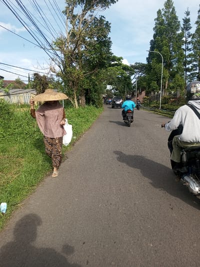 An individual with a straw hat walks on a narrow paved road bordered by a grassy verge. The person wears a brown top and patterned trousers, carrying a white bag. Behind them, two motorcyclists travel on the road, followed by a black car. Tall trees line the opposite side of the road, obscuring buildings. Overhead, power lines crisscross the sky under a partly cloudy sky.