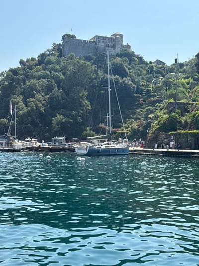 The image shows a body of water in the foreground, with several white buoys floating on the surface. In the middle ground, a harbor is visible with several boats docked, including a large sailboat with a blue hull. A stone pier runs along the waterfront, with a few people walking on it. Behind the harbor, a lush green hillside rises steeply, covered in trees and vegetation. At the top of the hill, a stone castle with red-tiled roofs is perched, overlooking the bay. The sky is clear and blue. This scene is characteristic of the picturesque harbor of Portofino, Italy, with its historic castle and vibrant waterfront.
