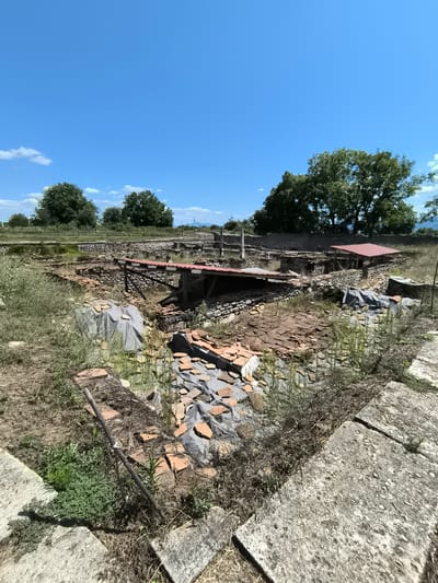 The image depicts the archaeological site of the ancient Roman villa in Garmen, Bulgaria. The foreground features weathered stone slabs and overgrown vegetation. In the midground, the excavated ruins of the villa are visible, characterized by stone walls, remnants of tiled floors, and areas covered with protective tarpaulins. Red-tiled roofs have been erected over some sections of the ruins to provide shelter. In the background, more stone structures and walls extend into the distance, bordered by a wooden fence. Lush green trees and a clear blue sky with scattered white clouds complete the scene. There are no people or animals visible in the image. The overall impression is one of an active archaeological excavation site under a bright, sunny day.