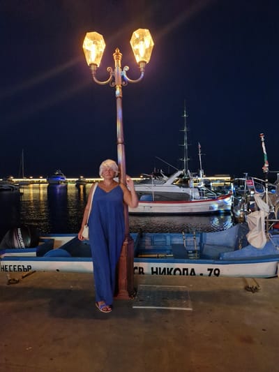 The image shows a woman standing next to a lamppost in a marina at night. She is wearing a blue jumpsuit and sandals, with a purse slung over her shoulder. The lamppost has two diamond-shaped lights. In the background, several boats are docked on the water, which reflects the lights of the marina. The marina is located in Sveti Vlas, Bulgaria. The boats are of various sizes, including a white yacht with red trim and a small blue fishing boat with the text "MERCURY" visible on an outboard motor and Cyrillic text on its side. A ladder is visible on the blue fishing boat. The ground appears to be concrete, with a metal grate set into it.