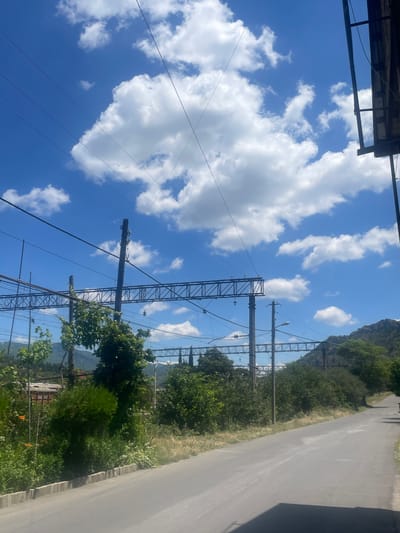 The image depicts a bright, sunny day in Mtskheta, Georgia, with a clear blue sky dotted with fluffy white clouds. The scene is dominated by a power line pylon and overhead wires, stretching across the frame. These lines traverse through lush green trees and vegetation that line a paved road, which occupies the right side of the image. In the background, a large, rocky hill is visible, partially covered by greenery. The perspective is upward-looking, emphasizing the vastness of the sky and the scale of the infrastructure against the natural landscape. The overall impression is one of a tranquil, rural or semi-rural environment, possibly near a railway line, given the presence of the power lines. There is no visible text in the image.