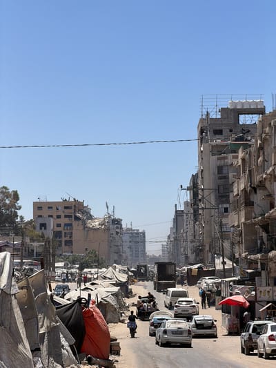 This is a vertical, eye-level, outdoor photograph captured on a sunny day, depicting a street scene in Gaza City, Palestinian Territories. The street is lined with damaged and partially destroyed buildings on both sides, indicating recent conflict or destruction. On the left side of the street, a collection of tents and makeshift shelters are set up, with various materials like white tarps, an orange tarp, and black fabric visible. In the foreground, a person on a scooter with a white box attached to the back is moving away from the camera on the right side of the street. Several cars are parked or driving on the street, including white sedans and a dark-colored van. Some of these cars appear to have damage or debris on them. In the background, the damaged buildings show exposed rebar, missing walls, and rubble. In the distance, more buildings can be seen, some of which appear to be under construction or repair with scaffolding. The sky is clear and blue. There are power lines and utility poles visible throughout the scene. The overall atmosphere suggests a scene of recovery or displacement amidst urban destruction.