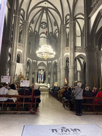 The image shows the interior of the Templo de Arucas, a church located in the city of Arucas, Gran Canaria, Spain (latitude: 28.119, longitude: -15.523). The church's interior is dominated by tall grey columns, intricate arches, and a large chandelier hanging from the high vaulted ceiling. The floor is tiled and there are several rows of pews filled with people attending a service. At the far end of the church, a small group of people are standing in front of an ornate altar decorated with candles. There are several people standing at the front, including two people in white robes. The church is decorated with Christmas trees and lights, and the walls are decorated with various religious artwork.