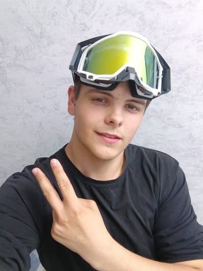 A young man poses for a selfie in Andreyevo, Russia. He wears a black T-shirt and ski goggles on his forehead. He's making a peace sign with his left hand. The background is a gray wall.