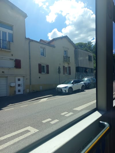 The image captures a street view in Nancy, France, likely from the perspective of a moving vehicle. The scene is dominated by residential buildings and a street with clear lane markings.
In the foreground, a modern white car is parked on the right side of the road. Behind it, a dark-colored SUV is also visible. The buildings lining the street are typical European style, with cream-colored facades, shutters, and windows. One building has a distinctive modern extension with dark windows. The sky above is bright blue with fluffy white clouds, suggesting a clear and sunny day, likely during daylight hours.
The overall impression is that of a quiet, urban residential area. The image is taken from an angle that includes a portion of the interior of what appears to be a bus or train, with a yellow-and-gray handrail visible on the right side of the frame. Faint text is visible on the white panel of the vehicle's interior, possibly indicating reserved seating or rules. The perspective gives a sense of observing the outside world from a sheltered viewpoint.