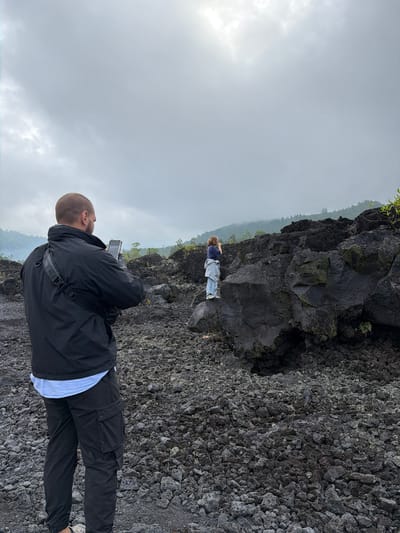 In Kintamani, Indonesia, two individuals are present in a rocky, volcanic landscape. In the foreground, a bald man wearing a black jacket, a light blue shirt, and black cargo pants is holding a smartphone to take a picture. He is positioned with his back to the camera, looking towards a person standing on a dark, porous rock formation in the mid-ground. This second person, seen from behind, is wearing a blue shirt, a grey jacket tied around their waist, and light-colored pants. They are also holding a phone, presumably taking a photo or video of the surrounding environment. The ground is covered in dark, irregular volcanic rocks and stones, with sparse vegetation visible on the rocky slopes and in the background. The sky is overcast with a thick layer of clouds, and a misty haze obscures the distant hills and trees. The overall scene suggests a visit to a volcanic site, possibly an active or recently active one, given the stark, rocky terrain.