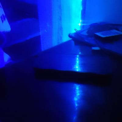 The image presents a dimly lit indoor scene, predominantly illuminated by vibrant artificial blue light. A smooth, dark, and highly reflective surface, resembling a table or desk, dominates the foreground and mid-ground. Bright, distinct points of blue light, consistent with LED strips, are reflected prominently on this surface, indicating a light source either above or along its edges.
In the mid-ground, resting on the reflective surface, is a dark, indistinct object, possibly a book, device, or a stack of items. A small, square-shaped label or sticker, featuring an unreadable pattern suggestive of a QR code or barcode, is visible on its upper side.
The background shows areas of intense blue and cyan illumination. A bright blue light illuminates a vertical structure on the left, possibly a wall. Another distinct light source, appearing more cyan, is visible in the upper right portion of the frame. The strong artificial lighting suggests the scene is set at night or in a windowless room. No people are present, and no specific activity is taking place. The visual information does not provide any specific location cues related to Nkaimurunya ward, Kenya.