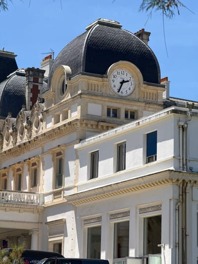 A close-up, eye-level, outdoor, daytime shot focuses on the upper floors and roof of a white and cream-colored building with architectural details. The building features multiple arched windows with decorative surrounds, a white balustrade along a balcony, and a large domed roof covered in dark gray tiles. A round clock with black numbers and hands is prominently displayed on the building's facade, beneath the dome. The sky is clear blue. The building appears to be a historic structure, possibly a hotel or public building. There is a hint of a tree branch in the upper right corner. The image appears to have been taken in Biarritz, France, a coastal resort town known for its Belle Époque architecture.