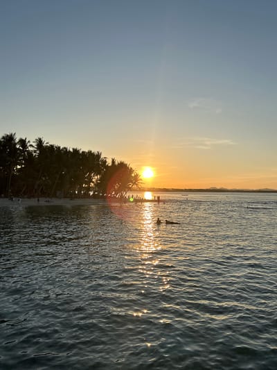 The image depicts a sunrise over the ocean at General Luna, Philippines. A row of palm trees lines the beach. People are standing in the water and on the beach. The sun reflects off the water's surface.