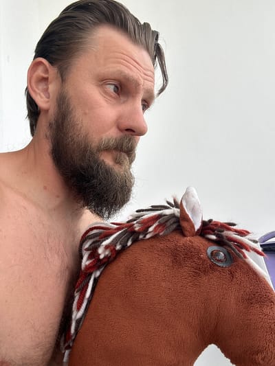 A close-up, waist-up shot shows a shirtless man with a beard and dark hair looking to his right, away from the camera. He is holding a stuffed toy horse, the head of which is visible in the foreground. The toy horse is brown with a multi-colored yarn mane. The background is a plain white wall. The image was taken in Vsevolozhsk, Russia.