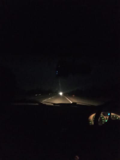 The image captures a nighttime view from inside a vehicle traveling on a highway. The dashboard lights, which appear to be green, are illuminated. The road ahead is visible with lane markings, and other vehicles' headlights and taillights can be seen in the distance. The overall scene is dark, suggesting a rural or suburban area outside the city of Almaty, Kazakhstan. There are no people clearly visible within the frame.