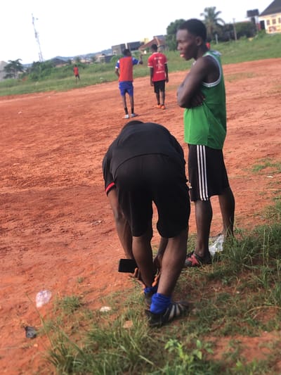 This image, taken in Nsugbe, Nigeria, captures an outdoor scene on a reddish-brown dirt field with patches of green grass. The sky is bright, indicating clear weather, likely midday or afternoon.

In the foreground, two individuals are prominent. On the left, a person with dark skin wears a bright green tank top and black shorts with white stripes down the side, standing with arms crossed and looking to their left. To their right, another person, also with dark skin, wears a black t-shirt and black shorts. This individual is bent over, seemingly stretching or adjusting something on their foot, while holding a black mobile phone in their left hand. They wear blue socks or wraps on their ankles, and dark footwear. Scattered on the grassy patches are pieces of plastic debris, including bags and bottles.

In the midground and background, other figures are visible. One person wears a red shirt and black shorts, standing further back. Another person in a red shirt and blue shorts is visible on the far left. The overall activity appears to be an informal sports practice or gathering. The individuals are dressed in athletic attire, suggesting a focus on physical activity on the open field.

The background shows sparse vegetation, including what appears to be a palm tree, and a few light-colored buildings. A tall structure resembling a power line tower is also faintly visible in the distance. No discernible text is visible on any clothing or objects. The atmosphere appears casual, possibly during a break or warm-up session.