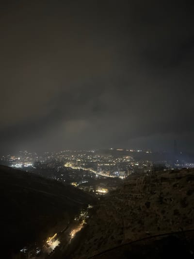 An aerial nightscape unfolds, dominated by dark, ominous clouds covering the left half. The right side reveals a steep, rugged cliff face framing an illuminated cityscape. Numerous lights form a winding pattern, suggesting a network of roads and buildings stretching into the distance. The terrain below appears mountainous, marked by dark shadows and sparse illumination, indicating a challenging landscape. A thin wire extends from the top right, adding a sense of height and perspective.