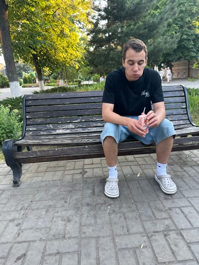 A young man is sitting on a dark wooden park bench, holding a small pink carton of juice with a straw. He is wearing a black t-shirt, denim shorts, white socks, and grey Crocs. His eyes are closed, and his mouth is slightly open, suggesting he is either about to drink or is enjoying the juice. The bench is made of dark wooden planks and has black metal legs, set on a grey paved walkway.
Behind the bench, lush green trees and bushes are visible. In the background, a few other people can be seen walking, some of whom appear to be children. The lighting suggests it is daytime, possibly late afternoon or early evening, with warm sunlight filtering through the trees. The setting appears to be a park or a public garden, likely in Azov, Russia. The overall mood is relaxed and casual. There is no visible text on the juice carton that can be read clearly.