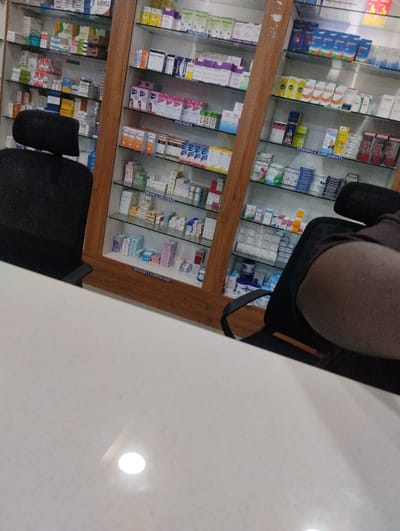 A pharmacy interior with a counter in the foreground. Shelves behind the counter are stocked with a variety of medications. Two black chairs are positioned near the counter, one appearing to be occupied. The shelves are organized with different drugs, vitamins, and supplements. The scene is well-lit, reflecting off the glossy counter surface.