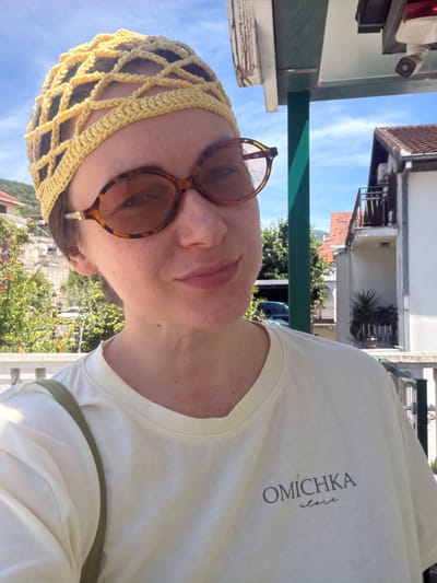 A person wearing sunglasses and a yellow crocheted head covering is shown from a low angle. The person is wearing a light yellow t-shirt with the inscription "OMICHKA slave" in dark gray lettering. They are standing outdoors, under a green awning. In the background, there are buildings and trees, suggesting the location is Meljine, Montenegro.