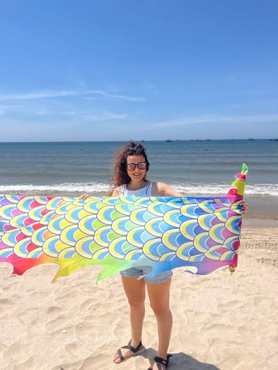 A young woman with curly dark hair and glasses stands on a sandy beach in Mui Ne, Vietnam. She is holding up a colorful, patterned fabric that resembles dragon scales, with shades of red, orange, yellow, blue, and pink. The fabric has a flowing, serpentine shape. The woman wears a white tank top and denim shorts. Behind her, the ocean stretches to the horizon under a clear blue sky with a few wispy clouds. Several small boats are visible on the water. The sand is light brown and textured with ripples and scattered shells. Her feet are clad in black sandals.