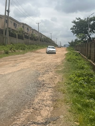 The image depicts a dirt road in Gwarinpa, Nigeria, bordered by a line of residential buildings on the left and a concrete block wall and vegetation on the right. A white sedan is parked on the left side of the road, facing away from the camera. Further down the road, another car is visible, and a person is walking in the distance. The sky is overcast with clouds. The road surface is uneven and appears to be unpaved, with patches of gravel and dirt. Lush green vegetation grows along the sides of the road and behind the wall on the right. The architecture of the houses suggests a modern residential development. The overall scene captures a typical streetscape in a suburban area.