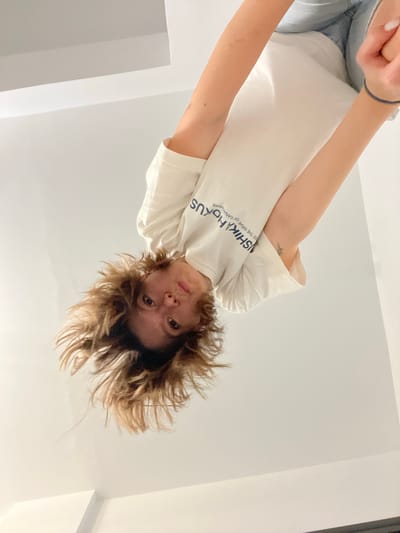The image shows a person with light brown, tousled hair, looking directly at the camera. They are wearing a white t-shirt with blue text that appears to read "ISHIKA HOKUS" and "ER THE WAVE OF KANAGAWA". The person is upside down in the frame, and their arms are visible. One arm is bent, and the hand is near their face. The other arm is extended downwards, and a dark blue bracelet is visible on their wrist. The background is a plain, light-colored wall or ceiling, with some architectural lines suggesting an indoor space. The lighting is bright and diffused. There are no discernible landmarks or clues to place this image in Limassol, Cyprus.