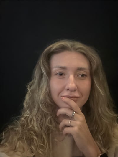 A young woman with blonde, wavy hair is the subject of this close-up portrait. Her face is angled towards the camera, with one hand touching her chin. She is wearing two rings on her right hand, one with a subtle detail. The lighting creates a gentle glow on her skin, highlighting her features. Her expression appears relaxed and contemplative. The background is a dark, solid color, which makes her stand out. The image is oriented sideways. There is no visible text or external cues to indicate the time of day, weather, or specific location within Chalong, Thailand. The overall mood is intimate and personal.