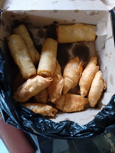 A close-up, high-angle shot captures a collection of fried snacks inside a cardboard box. The box, partially lined with a crumpled black plastic bag on the left, contains an assortment of golden-brown, crispy-looking items.

There appear to be two main types of snacks. On the left and top, several elongated, cylindrical items, resembling spring rolls or "lumpia" commonly found in Indonesia, are visible. They have a smooth, golden-brown wrapper. Scattered among these, and more prominent on the right side of the box, are smaller, irregularly shaped, or crescent-like fried pastries. These items suggest a variety of traditional Indonesian fried snacks, such as "pisang molen" (fried banana pastry) or other types of fried dumplings.

The cardboard lining of the box shows noticeable oil stains from the fried food, indicating their freshly prepared and oily nature. The snacks are piled somewhat haphazardly, suggesting they are either freshly placed or have settled during transport. A glimpse of a brown leather object, possibly a wallet or bag, is visible in the bottom-left corner outside the box. The lighting suggests an indoor setting, but the time of day and specific location within Ciamis, Indonesia, are not discernible. No people or text are visible in the image.