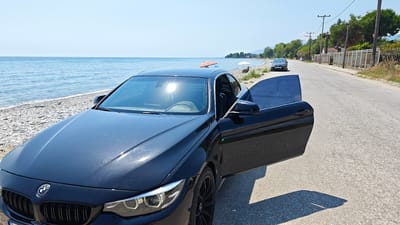 This media file captures a glossy black BMW coupe parked on the side of a road next to a pebble beach. The car's driver-side door is open, revealing a glimpse of the interior. The scene is set on a sunny day, with the bright sun reflecting off the car's windshield and the calm, blue ocean stretching out to the horizon.

In the background, several beach umbrellas are scattered along the sandy shore, with a few people visible enjoying the beach. A woman sits under a white umbrella, and another individual is partially visible near a striped umbrella. Further down the road, another car is parked. The road is asphalt and appears to be a coastal route. Telephone poles and lush green trees line the opposite side of the road, with some buildings and a white fence in the distance.

The time of day appears to be midday, given the strong sunlight and the shadows cast. The weather is clear and warm, characteristic of a summer day at the beach. The location cues, such as the Mediterranean-style vegetation and the overall coastal setting, are consistent with Leptokarya, Greece. The image evokes a sense of leisure and relaxation, with the car appearing to be a focal point of a holiday or excursion. No visible text is present in the media file.
