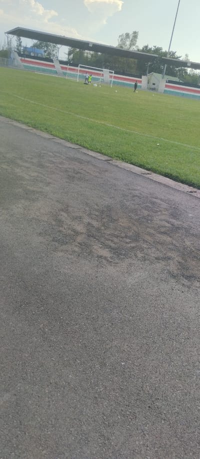 The image shows a paved pathway beside a soccer field in Kisumu, Kenya. The field is covered in short green grass, bordered by a white line. There are goal posts and a few players in the distance. There are trees lining the field in the background. The sky is mostly clear, with some scattered clouds. The pathway is made of asphalt, which is covered in dust and dirt. There is a concrete curb separating the path from the field.