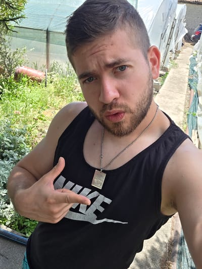 A man with short, dark hair and a beard is wearing a black tank top with white lettering, and a silver chain with a pendant. He is making a gesture with his left hand. The background includes greenery and what appears to be a partial view of a building or structure covered in white material. The location is Yagodovo, Bulgaria.