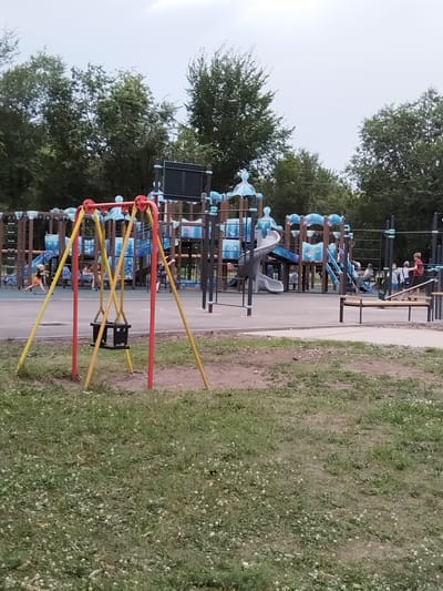 The image depicts a playground in Рощинский, Russia. In the foreground, a swing set with two black seats is visible. The playground equipment is a multi-colored structure with slides, stairs, and platforms. Several children and adults are present on the playground, engaged in various activities. Some children are playing on the equipment, while others are walking or standing around. The ground of the playground is paved with asphalt, and the surrounding area is covered with grass and trees. The sky is overcast.