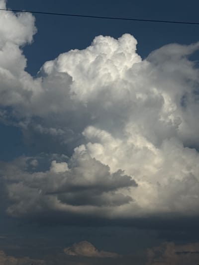 A vertical, slightly low-angle, eye-level medium shot captures a dynamic sky filled with dramatic clouds. The sky is a deep, rich blue, hinting at a late afternoon or early evening setting. A thin, dark horizontal line, likely a power line, stretches across the top of the frame. The clouds are the dominant subject, exhibiting a wide range of tones and textures. On the left and center, large, billowing cumulus clouds are illuminated from above, appearing bright white with soft, rounded edges. These clouds have a sense of depth and volume, with shadowed undersides that create contrast and highlight their three-dimensional form. Moving downwards and towards the right, the clouds transition into darker, more ominous cumulonimbus formations. These lower clouds are a deep gray, appearing heavy and dense, suggesting the potential for precipitation. The contrast between the bright, sunlit cumulus clouds and the dark, shadowed cumulonimbus clouds creates a visually striking and atmospheric scene. The overall impression is one of an impending storm or a dramatic shift in weather. The image does not contain any people, discernible structures or landmarks that would specifically identify the location as Bessonovka, Russia.