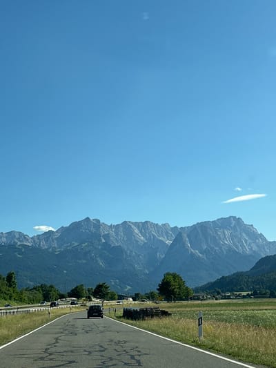 This is a landscape image taken from a moving vehicle on a road in Farchant, Germany. The road is asphalt and has white lines marking the lanes. There is a black car driving ahead of the viewer on the road. To the left of the road, there is a highway with several cars on it. A grassy field with tall yellow grass is on the right side of the road. In the background, there are green trees and buildings. Behind the trees and buildings, a large mountain range with rocky peaks dominates the horizon. The sky is clear and blue with a few wispy clouds.