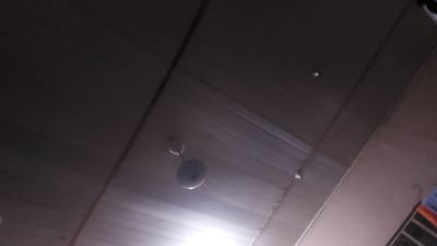 The image shows a ceiling fan mounted on a wooden plank ceiling. The fan is light-colored and appears to be a standard ceiling fan with a central motor housing and blades. The ceiling is made of parallel wooden planks, some of which are illuminated by a bright light source, possibly a bulb within the fan or a separate light fixture. The scene is dimly lit overall, with shadows cast by the fan and the ceiling structure. There is a small, circular light fixture or sensor visible on the ceiling in the upper right quadrant of the image. A sliver of what appears to be a colorful fabric or material is visible in the far right edge of the image. The perspective is from below, looking upwards. The image is assumed to be from New Nyanya, Nigeria, based on the provided context.