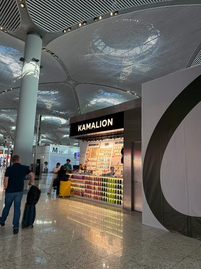 The image depicts an interior view of a modern airport terminal in Arnavutköy, Turkey. A man in a dark t-shirt and jeans is seen from behind, pulling two suitcases, one black and one dark green, across a polished granite floor that reflects the overhead lighting and the storefronts. To his right is a brightly lit kiosk named "KAMALION" which displays an array of accessories, including phone cases, jewelry, and electronic gadgets. Behind the kiosk, a few individuals, one wearing a green shirt, are visible interacting. The terminal features a distinctive architectural design with a high, slatted ceiling supported by large, cylindrical columns. A large, partially visible white banner with a dark curved stripe is positioned to the right of the kiosk, suggesting an advertisement or display. The overall environment suggests a bustling commercial area within an airport.